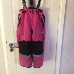 Termobyxor från t.sportswear - Rosa termobyxor från t.sportswear med hängslen, reflexdetaljer och flera fickor. Byxorna har förstärkta knän och dragkedjor vid fickorna. Bra skick och storlek 116
