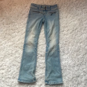 Ljusblå bootcut jeans med dragkedjor - Snygga ljusblå jeans med bootcut passform och låg midja. Jeansen har slitningar framtill och coola dragkedjor vid fickorna. Perfekta för dig som gillar en retro vibe och vill sticka ut lite extra. Klassisk femficksmodell och sköna att bära hela dagen.