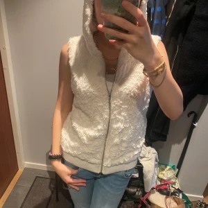 Vit fluffig pälsväst med luva - Mysig vit pälsväst med luva och dragkedja framtill. Västen är fluffig och har ribbade muddar nedtill för en skön passform. Perfekt att slänga över en hoodie eller t-shirt för en chill look.