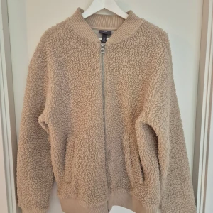 Beige teddyjacka från H&M, M - Mysig beige teddyjacka från H&M i relaxed fit. Jackan har dragkedja framtill, ribbade muddar vid ärmslut och nederkant samt två fickor. Perfekt för kalla dagar när du vill vara både varm och snygg.