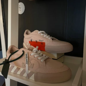 Off-White rosa sneakers med ränder - Säljer ett par Off-White sneakers i ljusrosa canvas med vita snören och ikoniska Off-White-detaljer som texten 'SHOELACES' på snörena, randig sula och orange tag. Snygg låg modell med rund tå och platt sula. Kommer med originalkartong och dustbag.