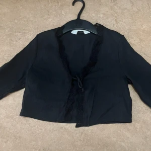 Svart bolerotopp med volangärm H&M - Svart bolerotopp från H&M med knyt framtill och dekorativ spetskant längs urringningen. Toppen har långa ärmar med volangdetalj vid ärmslut och är tillverkad i ett lätt, något glansigt tyg. Perfekt för att lyfta en outfit med en cool och chic touch.