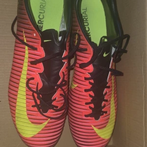 Nike Mercurial fotbollsskor röd/gul storlek 43 - Säljer ett par Nike Mercurial fotbollsskor i rött med svarta och gula detaljer. Skorna har snörning, svart plös och gula dobbar. Snyggt randigt mönster och stor Nike-logga på sidan. Perfekta för dig som vill sticka ut på planen.
