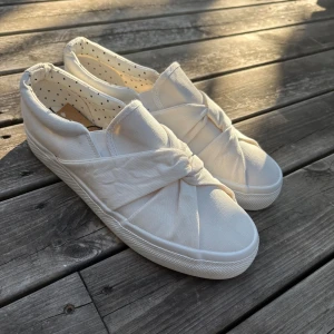 Vit skor - Säljer ett par vita slip-on sneakers i tyg med snygg knutdetalj framtill. Aldrig använda och perfekta till exempelvis student💕