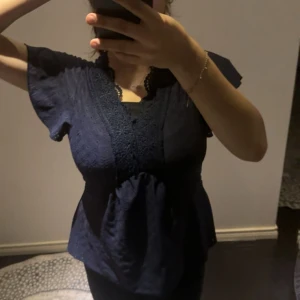 Mörkblå blus med spetsdetaljer - Supersöt mörkblå blus med v-ringning och spetsdetaljer längs halsringningen. Blusen har korta ärmar och lätt utställd passform nedtill. Perfekt för dig som gillar romantiska och feminina detaljer.