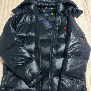  Polo Ralph Lauren jacka - Snygg svart pufferjacka från Polo Ralph Lauren med glansig finish och röd logga på bröstet. Jackan har huva, flera dragkedjor och quiltad design. Perfekt för kalla dagar och har en modern, oversized vibe.