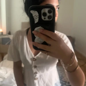 Vit blus med knappar och v-ringning - Säljer en vit blus med korta ärmar och snygg v-ringning. Blusen har fyra stora, bruna knappar framtill och en lätt figurnära passform. Materialet känns tunt och luftigt, perfekt för varmare dagar. Enkel och stilren design som är lätt att matcha. Storlek är s och märket är Gina 