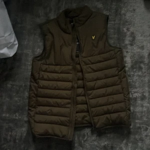 Brun/grön  väst från Lyle & Scott - Snygg brun dunväst från Lyle & Scott med klassisk gul logga på bröstet. Västen har quiltad design, hög krage och dragkedja framtill. Perfekt att slänga över en hoodie för extra värme. Enkel och stilren look som funkar till det mesta.