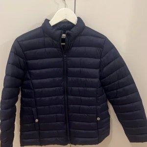 Mörkblå pufferjacka från MNG - Snygg mörkblå pufferjacka från MNG med horisontella sömmar och dragkedja framtill. Jackan har två fickor med tryckknappar och står upp lite i kragen.