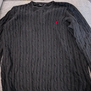 Svart kabelstickad tröja med röd logo stl 146-152 - Svart kabelstickad tröja från Mywear med rund halsringning och en röd broderad hjort på bröstet. Tröjan har lång ärm och ribbade muddar vid hals, ärmslut och nederkant. Perfekt för dig som gillar klassisk stil med en twist.