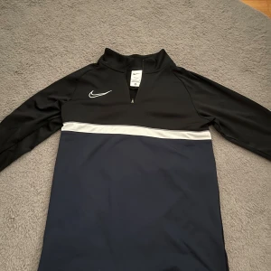Nike Dri-Fit svart/blå half zip tröja - Nike Dri-Fit tröja med svart överdel, mörkblå nederdel och vit rand över bröstet. Långärmad modell med half zip och broderad vit swoosh på bröstet. Perfekt för träning eller chill dagar. Materialet är polyester som andas och känns lätt på kroppen.