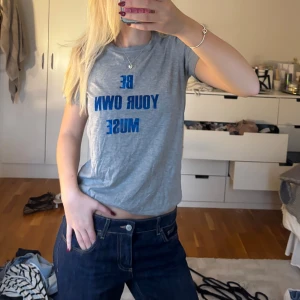 Grå t-shirt - Grå t-shirt i bomull med blå texten 'BE YOUR OWN MUSE' framtill. Klassisk passform med korta ärmar och rund halsringning. Perfekt för en chill och avslappnad stil.