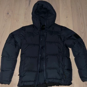 Marinblå peak performance puffer M - Marinblå pufferjacka från Peak Performance med huva och dragkedja. Jackan har en quiltad design, två sidofickor med dragkedja och en diskret logga på ärmen. Perfekt för kalla dagar och riktigt skön att bära, säljer för att jag har vuxit ur den. 