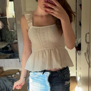 Vit blus med volang och knyt i ryggen - Supersöt vit blus med broderade detaljer, volang vid axlarna och peplum-aktig nederdel. Toppen har fyrkantig ringning och öppen rygg med knytband. Perfekt till jeansshorts för en somrig look!