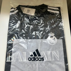 Real Madrid x Balmain x Adidas fotbollströja - Exklusiv svart fotbollströja från Real Madrid i samarbete med Balmain och Adidas. Tröjan har korta ärmar, vita detaljer på axlarna och Real Madrid-loggan på bröstet. Materialet är lätt polyester med Aeroready-teknologi för bästa komfort.