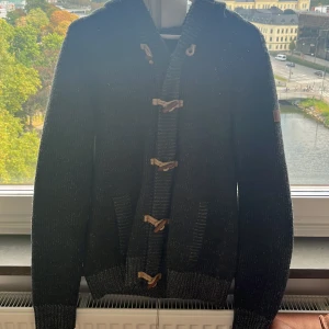 Svart stickad kofta från Petrol Industries - Svart stickad kofta från Petrol Industries med grov ribbad krage och snygga träknappar i snörning längs framsidan. Koftan har två fickor framtill och långa ärmar, perfekt för kyliga dagar. Materialet är mjukt och värmande stickat.