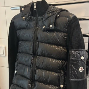 Moncler Cardigan bra skick! - Säljer en svart dunväst från Moncler med glansig finish, huva och stickade ärmar. Västen har dragkedja framtill, tryckknappar och en ikonisk Moncler-logga på ärmfickan. Perfekt för lager-på-lager och streetstyle. Storlek L men passar även M. Priset ej hugget