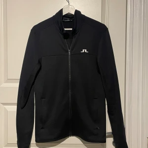 Svart ziptröja från J.Lindeberg - Svart långärmad ziptröja från J.Lindeberg med vit logga på bröstet. Tröjan har hel dragkedja framtill, ståkrage och två diskreta fickor. Materialet är mjukt och stretchigt, perfekt för en clean och sportig look.