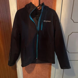 Svart vindjacka från Everest - Svart vindjacka från Everest med huva och blå detaljer vid dragkedjan. Jackan har lång ärm och en sportig look, perfekt för dig som gillar att vara aktiv utomhus. Snygg logga på bröstet och praktiska fickor med dragkedja.