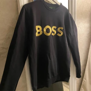 Svart sweatshirt från BOSS med guldtryck - Svart sweatshirt från BOSS med stort, glansigt guldtryck på bröstet. 