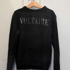 Svart sweatshirt Zadig & Voltaire - Cool svart sweatshirt från Zadig & Voltaire med silvrigt tryck VOLTAIRE på ryggen. Klassisk rund halsringning och långa ärmar. Perfekt för dig som gillar stilrena och trendiga plagg med en touch av streetstyle.