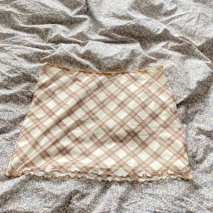 Beige och brun rutig minikjol - Supersöt kortkjol i beige och brunt rutigt mönster med volangkant och liten rosett i midjan. Kjolen har en lätt och luftig känsla, perfekt för dig som gillar en söt och trendig stil.