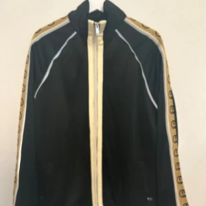 Svart Gucci vindjacka med logoband - Svart vindjacka från Gucci med beige logoband längs ärmarna och dragkedja framtill. Jackan har vita detaljer och hög krage. Tillverkad i 100% bomull, perfekt för dig som gillar exklusiv streetwear och vill sticka ut med designerplagg.