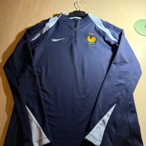 Frankrike Nike Dri-Fit fotbollströja XL - Säljer en marinblå långärmad Frankrike fotbollströja från Nike i Dri-Fit material. Tröjan har halv dragkedja, broderad tupp-logga med två stjärnor och vita detaljer längs ärmarna. Slim fit-modell, perfekt för träning eller match.