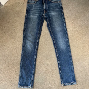 Blå jeans från Nudie Jeans, 29/32 - Snygga blå jeans från Nudie Jeans med klassisk femficksdesign och orangea sömmar. Jeansen har en rak passform och är tillverkade i slitstarkt denimtyg av bomull. Perfekta för dig som gillar en tidlös och avslappnad stil. Obs jeansen har ett lagat hål vid midjan men inget man tänker på!