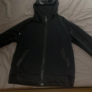 Svart hoodiejacka från C.P. Company - Svart hoodiejacka från C.P. Company med ikoniska goggles i huvan. Hoodien har dragkedja framtill, två fickor med dragkedja. Ena googglen är lös så därför säljer jag för billigare