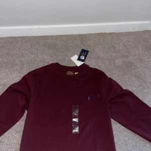 Vinröd långärmad tröja Polo Ralph Lauren - Vinröd långärmad tröja från Polo Ralph Lauren med klassisk rund halsringning och blå broderad logga på bröstet. Tillverkad i mjuk bomull, perfekt för en stilren och avslappnad look.