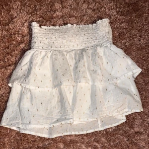 Vit volangkjol med smock i midjan - Supersöt vit kortkjol med volanger och smockad midja. Kjolen har små guldiga prickar i tyget och är gjord i ett lätt bomullsmaterial. Perfekt för varma dagar och ger en romantisk vibe.