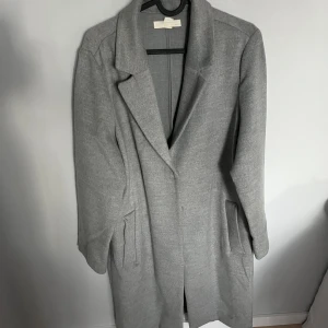 Grå kappa från H&M, storlek 38 - Snygg grå kappa från H&M i klassisk modell med krage och knäppning framtill. Kappan har två fickor på sidorna och är i ett mjukt material som känns både stilrent och bekvämt. Perfekt för dig som gillar en clean och tidlös look. Använd fåtal gånger och i mycket bra skick. Inga defekter eller fläckar förutom på lappen, syns på bild 4.