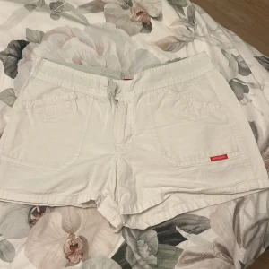 Vita shorts från Birdira, storlek L - Snygga vita shorts från Birdira i storlek L. De har en klassisk midwaist-passform, stora fickor fram och bak med knappdetalj, samt en diskret röd logotyp. Materialet känns som mjuk bomull, perfekt för varma dagar.