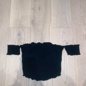 Svart offshoulder croppad topp - Trendig svart offshoulder topp med croppad passform och ribbad struktur. Toppen har korta ärmar som sitter under axlarna och volangkant både upptill och nedtill för en söt touch. Perfekt för dig som gillar en stilren men ändå lekfull look. Sista bilden är samma tröja i vit färg för att se hur modellen sitter.