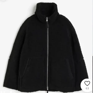 Svart teddyjacka med dragkedja - Mysig svart teddyjacka med hög krage och grov silverfärgad dragkedja. Jackan har två stora fickor framtill och en oversized passform som ger en chill vibe. Perfekt för kalla dagar när du vill vara både varm och snygg.