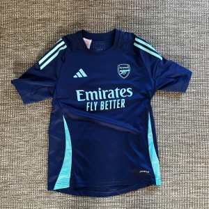 Arsenal blå fotbollströja Adidas - Snygg marinblå Arsenal fotbollströja från Adidas med ljusblå detaljer och logga. Tröjan har korta ärmar, rund hals och är tillverkad i lätt och ventilerande material (Aeroready). Perfekt för träning eller match!