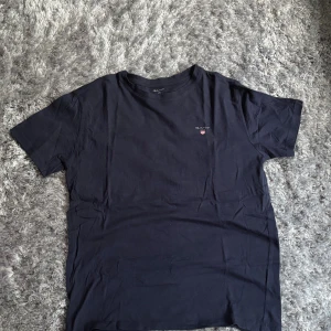 Svart t-shirt från Gant - En stilren svart t-shirt från Gant med liten broderad logga på bröstet. Klassisk passform och rund halsringning, perfekt till jeans eller shorts. Tillverkad i mjuk bomull för maximal komfort.