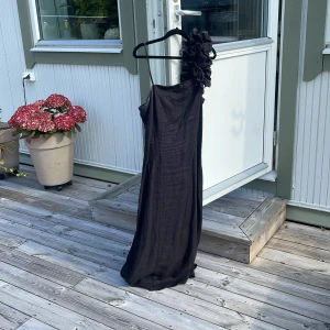 Svart långklänning med volangaxel - Säljer en svart långklänning med en axel och stor volangdetalj på axeln. Klänningen har en slits bak och är oneshoulder med smal axelrem på ena sidan. Materialet ser ut att vara linne eller liknande, vilket ger en snygg struktur.jag hade på mig den här klänning på min bal i 9an 2024