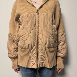 Beige quiltad cardigan jacka med dragkedja - Säljer en beige cardigan jacka med quiltade partier och stickade detaljer. Jackan har dragkedja framtill, ribbade muddar och krage samt en loose passform. Materialmix av stickat och syntet ger en unik look. Perfekt för hösten, lager-på-lager och chill stil