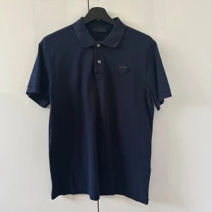 Mörkblå pikétröja från Prada - Snygg mörkblå pikétröja från Prada i klassisk modell med krage och två knappar framtill. Tröjan har diskret logga på bröstet och är tillverkad i mjuk bomull. Perfekt för en clean och stilren look. Endast använd 5 gånger och mycket bra skick. Skriv för fler bilder och frågor