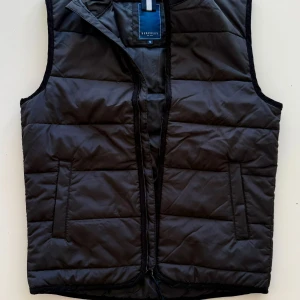 Svart dunväst från Berkeley - Snygg svart dunväst från Berkeley med quiltad design och dragkedja framtill. Västen har två fickor på sidorna och en klassisk, stilren look som passar perfekt över en hoodie eller tröja. Perfekt för kyliga dagar när du vill hålla värmen utan att tumma på stilen. Den passar även S bra. Och är helt ny oandvänd.