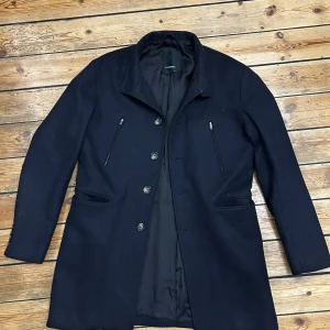 Svart rock från J.Lindeberg, stl 50 (M/L) - Stilren svart rock från J.Lindeberg med klassisk krage och knäppning framtill. Jackan har två bröstfickor med dragkedja, två sidofickor och dekorativa knappar vid ärmslut och baktill. Perfekt för dig som vill ha en clean och tidlös look.