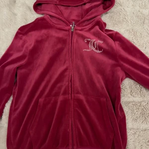 Rosa velour hoodie Juicy Couture - Juicy Couture hoodie i mörkrosa velour med glittrande strasstenar på bröstet och ryggen. Klassisk huva, dragkedja framtill och kängurufickor. Ikonisk logga i stenar både fram och bak. Perfekt för dig som älskar Y2K-stil!