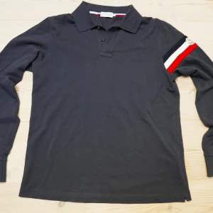 Svart långärmad pikétröja Moncler - Svart långärmad pikétröja från Moncler i slim fit den är M men passar också S. Tröjan har klassisk krage, knäppning framtill och Moncler-logga på ena ärmen. Ärmen har en snygg randig detalj i rött, vitt och blått. Tillverkad i mjuk bomull. Vid några frågor hör av er! Pris är ej hugget i sten! 