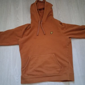 Brun hoodie från Lyle & Scott - Säljer en snygg brun hoodie från Lyle & Scott med klassisk gul logga på bröstet. Tröjan har huva med snörning, magficka och långa ärmar. Perfekt för chill dagar och enkel att matcha med jeans eller joggers.