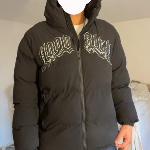 Svart pufferjacka från Hoodrich M - Svart pufferjacka från Hoodrich i storlek M. Jackan har stor vit logga över bröstet, dragkedja framtill och två sidofickor med dragkedja. Tjock och varm med huva, perfekt för kalla dagar. Snygg streetstil och riktigt bekväm passform. Nypris 1500