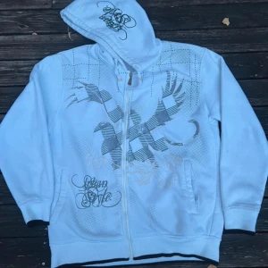 Vit hoodie med tryck från Sports - Vit hoodie med dragkedja och huva från Sports. Framsidan har ett stort grafiskt tryck i grått och vitt samt textdetaljer. Hoodien har fickor på sidorna och ribbade muddar vid ärmslut och nederkant. Perfekt för en chill och avslappnad stil. Tröjan är oversized!!