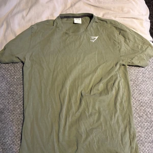 Olivgrön t-shirt från Gymshark - Olivgrön t-shirt från Gymshark med diskret logga på bröstet. Klassisk rund halsringning och korta ärmar. Perfekt för dig som gillar en enkel och sportig stil. Tillverkad i mjuk bomull som känns skön mot huden.