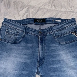 Replay Anbass jeans i ljusblå denim - Säljer ett par Replay Anbass jeans i ljusblå tvätt med klassisk femficksdesign och snygga slitningar. Jeansen har normal passform och raka ben, med detaljerade sömmar och logga på fickan. Perfekta för dig som gillar stilrena och bekväma jeans.